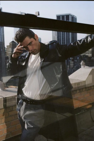 Man wearing a black leather trench coat over a white t-shirt and grey trousers, posing on a rooftop with city buildings behind.