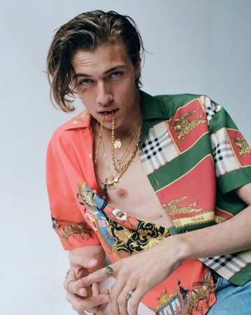 Young man with styled brown hair biting a gold chain wearing a patterned short sleeve shirt against a pale studio background.