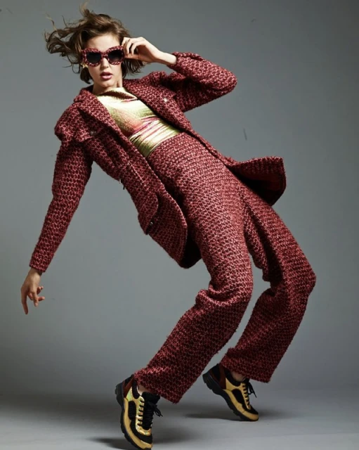 A woman models red tweed suit and shades, captured in full shot against a gray backdrop, showcasing an editorial fashion style.