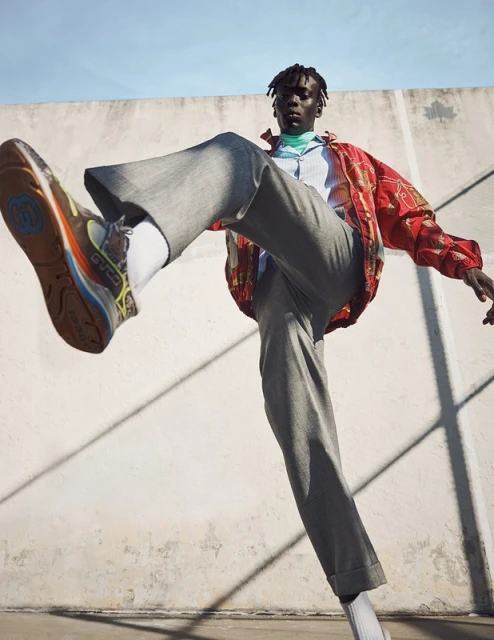 Fashionable African male model strikes dynamic pose outdoors in colorful shirt, grey wide-leg trousers and sneakers against weathered wall.
