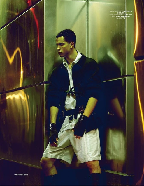A stylish adult male poses indoors in an elevator wearing a dark jacket, light shorts, shirt, fingerless gloves, and distinctive belt.