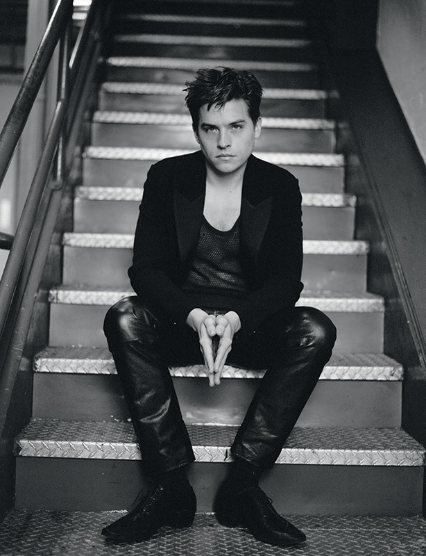 Monochrome portrait of a young adult man sitting on metal stairs, wearing a black suit jacket and leather pants, his hands clasped together.