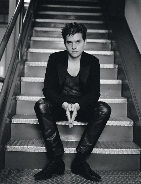 Monochrome portrait of a young adult man sitting on metal stairs, wearing a black suit jacket and leather pants, his hands clasped together.