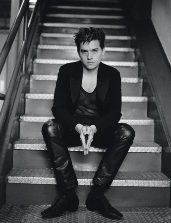 Monochrome portrait of a young adult man sitting on metal stairs, wearing a black suit jacket and leather pants, his hands clasped together.