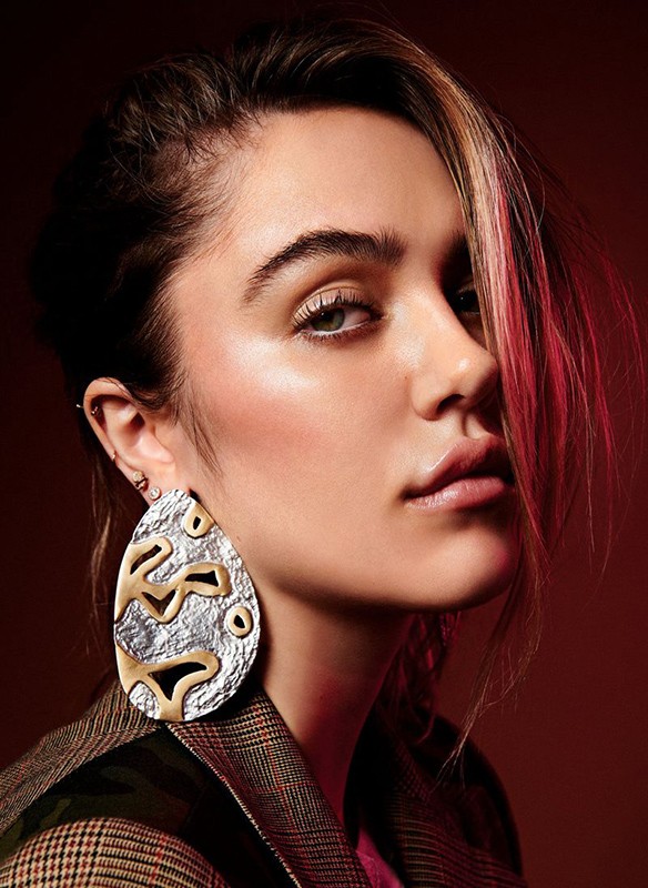 Portrait of a young adult woman in a studio, wearing large statement earrings, a patterned jacket, and pink shirt against a red backdrop.