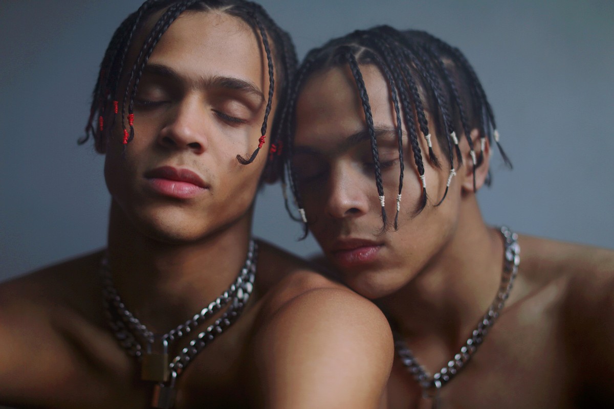Close-up portrait of two shirtless young adult males with braids and chains, close together against a grey backdrop in soft lighting.