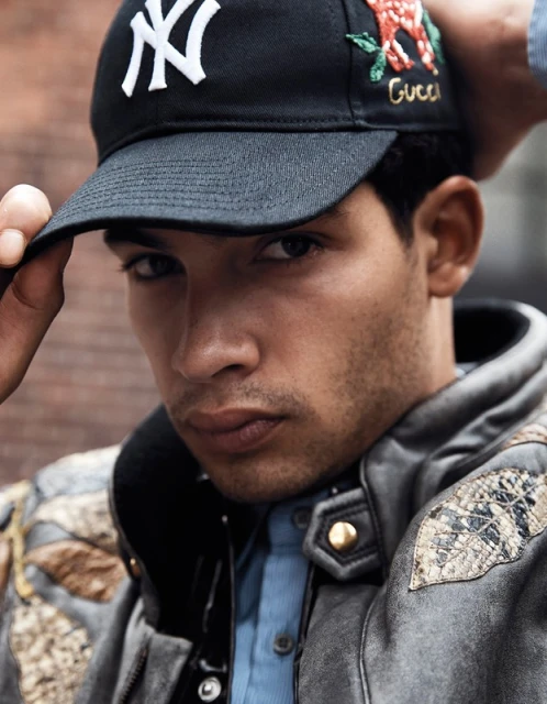 Close-up of a young adult male wearing an embroidered black NY baseball cap and patterned leather jacket, posing in an outdoor setting.