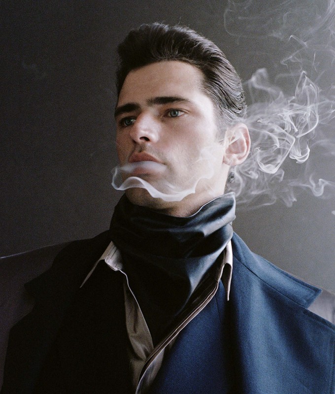 Portrait of a man with slicked-back dark hair, wearing a dark coat and turtleneck sweater, exhaling a plume of smoke against a dark backdrop.