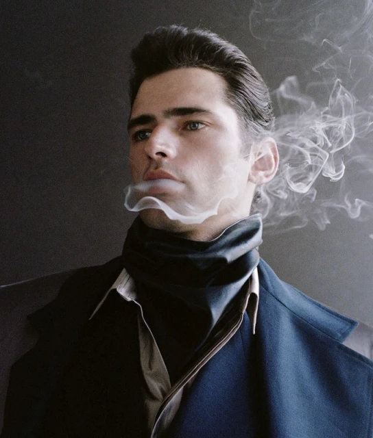 Portrait of a man with slicked-back dark hair, wearing a dark coat and turtleneck sweater, exhaling a plume of smoke against a dark backdrop.