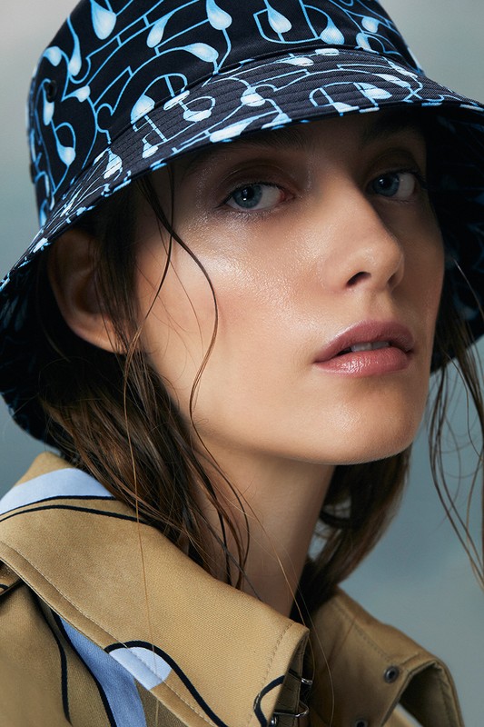 Close-up of a woman wearing a patterned bucket hat and tan jacket, showcasing fashion and beauty in a studio setting.