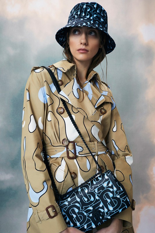 Woman in a patterned trench coat and bucket hat holding a matching handbag against a cloudy background in a fashion editorial shot.