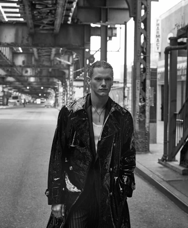 Black and white shot of a man in a shiny black coat standing on a city street under elevated train tracks, with a pharmacy sign visible.