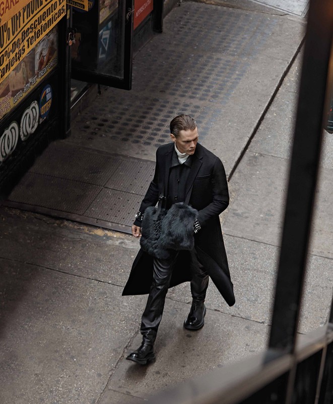 A stylish adult male in a long black coat walks on a city street. He carries a black fur bag and wears black leather pants and boots.
