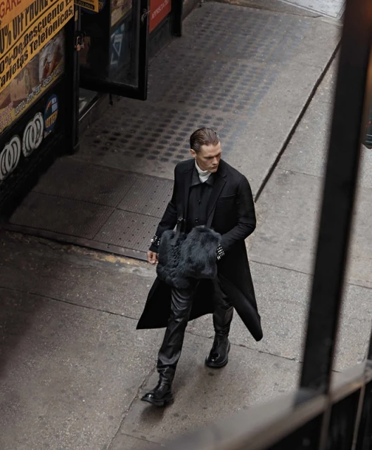 A stylish adult male in a long black coat walks on a city street. He carries a black fur bag and wears black leather pants and boots.