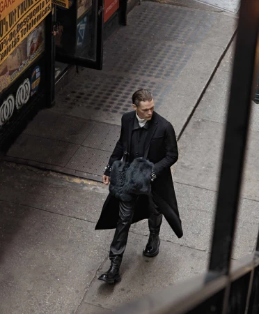A stylish adult male in a long black coat walks on a city street. He carries a black fur bag and wears black leather pants and boots.