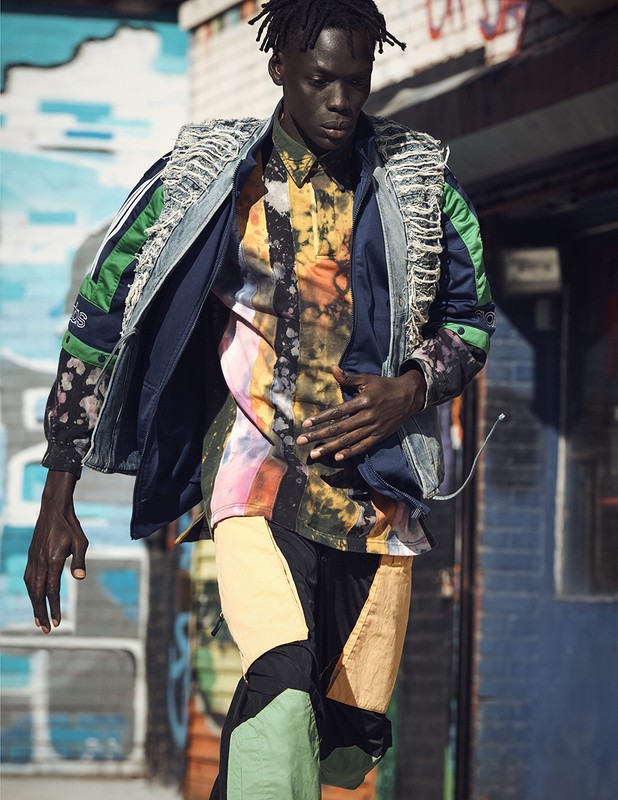Dark-skinned male model in colorful, deconstructed fashion attire against a graffiti wall background. Fashion editorial photography, outdoor setting.