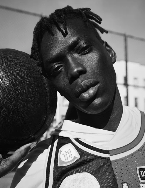 Black and white portrait of a male basketball player with dreadlocks leaning on a basketball on an outdoor court.