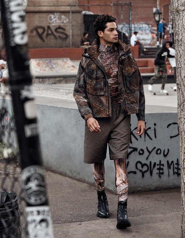 A fashionable young man in a patterned jacket and shorts strides confidently in an urban skatepark, blending style with the raw setting.