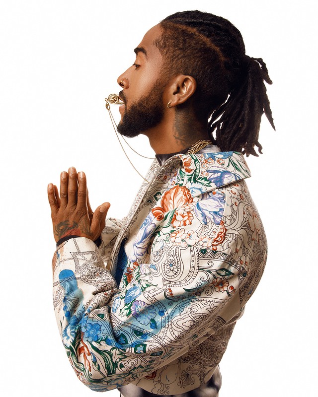 Profile portrait of Black man with dreadlocks and floral jacket posing in photo studio with hands clasped and ornate whistle necklace.
