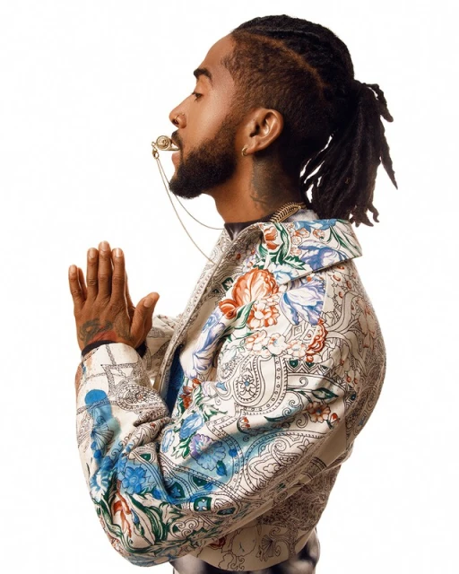 Profile portrait of Black man with dreadlocks and floral jacket posing in photo studio with hands clasped and ornate whistle necklace.