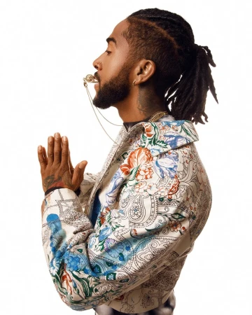 Profile portrait of Black man with dreadlocks and floral jacket posing in photo studio with hands clasped and ornate whistle necklace.
