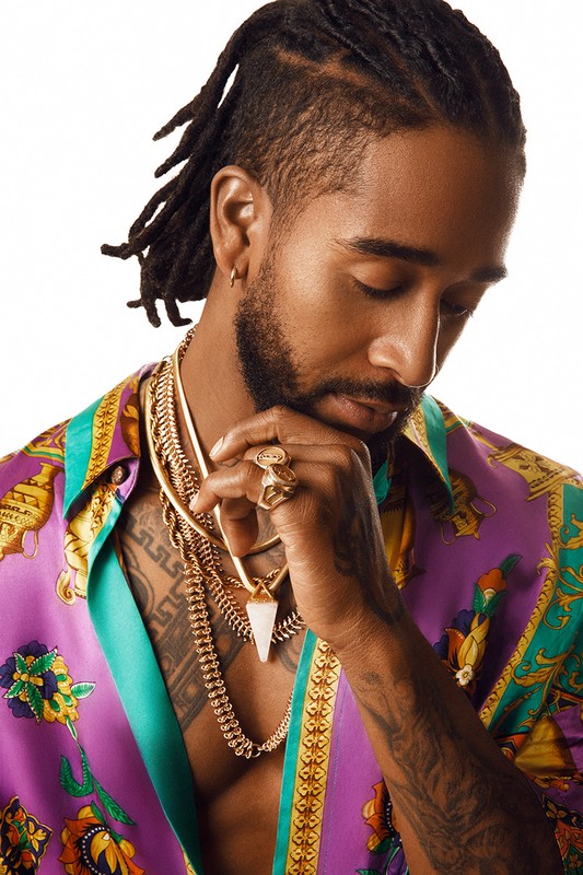 Portrait of an adult male with dreadlocks in a patterned shirt and jewelry standing against a white background in a studio setting.