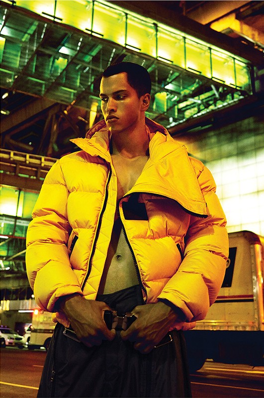 Fashionable adult male in a bright yellow puffer jacket poses outdoors at night against a cityscape and transit backdrop.