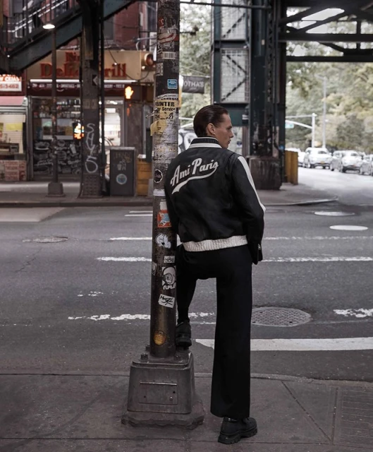A man in a black jacket with 'Ami Paris' on the back stands on a street corner, leaning against a lamppost in an urban setting with cars.