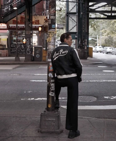 A man in a black jacket with 'Ami Paris' on the back stands on a street corner, leaning against a lamppost in an urban setting with cars.