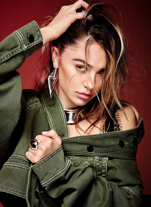 Portrait of a young adult woman with tan skin wearing a green denim jacket, jewelry, and dark eye makeup against a red background.