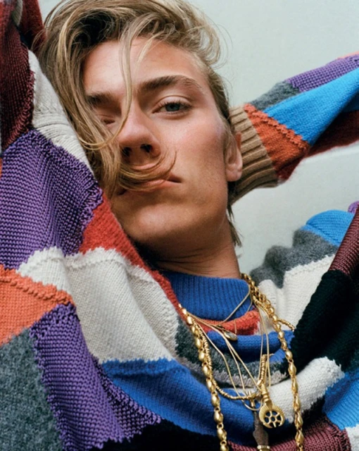 A young adult male poses indoors wearing a colourful knitted sweater and gold necklaces, looking towards the viewer with a relaxed expression.