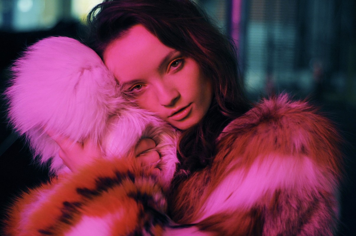 Portrait of a young woman with long brown hair, wearing a fur coat, holding a white fur muff. Pink and blue neon lighting.
