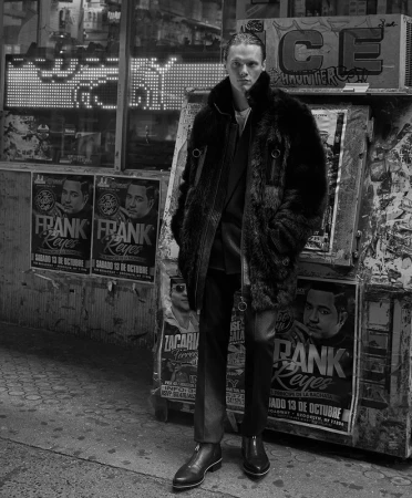 Man wearing a textured faux fur coat standing on a city street with posters and an illuminated sign in the background