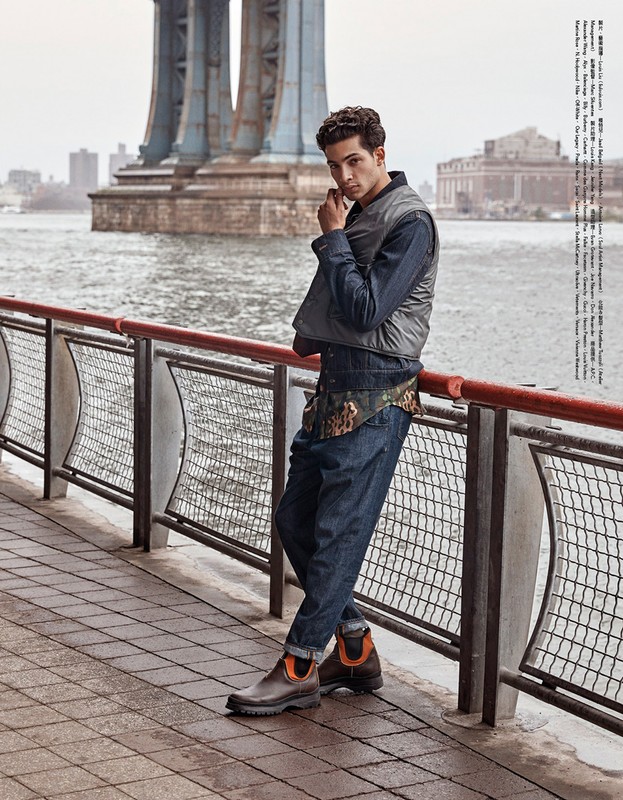 Adult male in fashion shot wearing denim shirt, vest, blue jeans, and brown boots leaning against a railing with bridge in background.