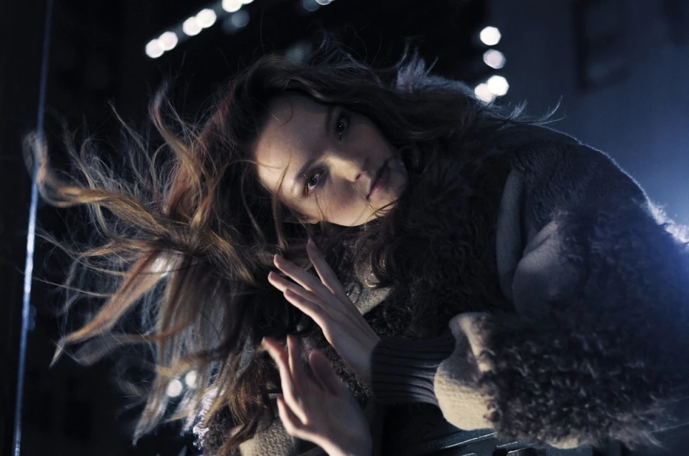 Portrait of a woman with wind-blown hair wearing a fur-trimmed jacket, looking directly at the camera against a dark background.