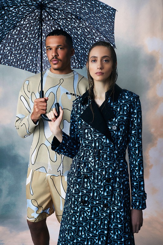 Two fashion models pose in matching patterned Burberry outfits, woman in a trench coat, man in a sweater and shorts, under an umbrella.