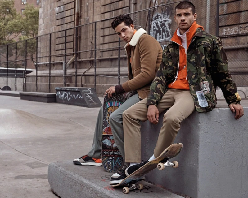 Two young adult men in urban skatepark. One holds skateboard, the other sits on ledge. Both wear jackets in an outdoor setting.