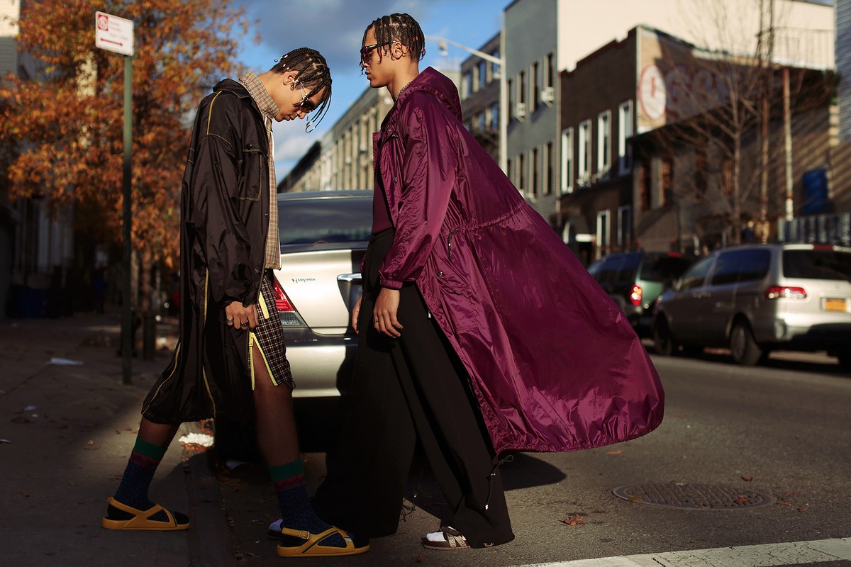 Two fashionable adults model long coats on a city street. One coat is burgundy, the other is black with yellow accents, daytime.
