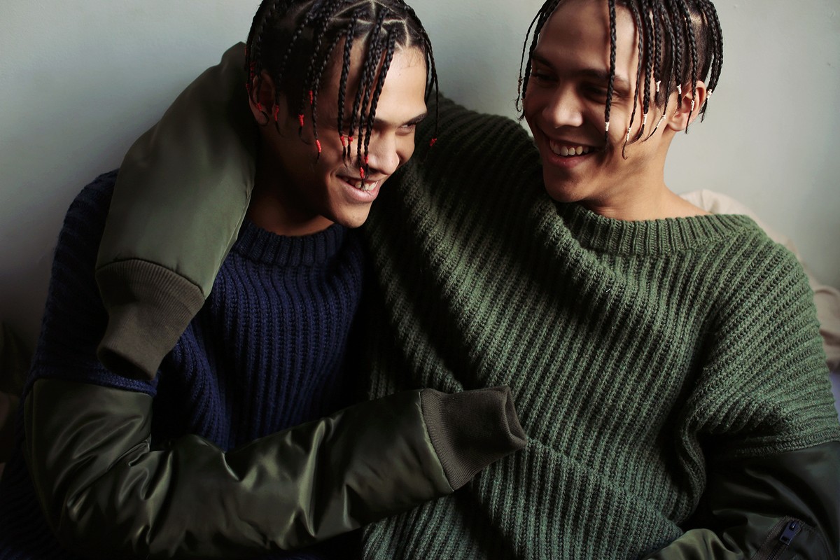 Two young adult men with braided hair smile in a close-up studio portrait, wearing knit sweaters with layered-sleeve bomber jackets.