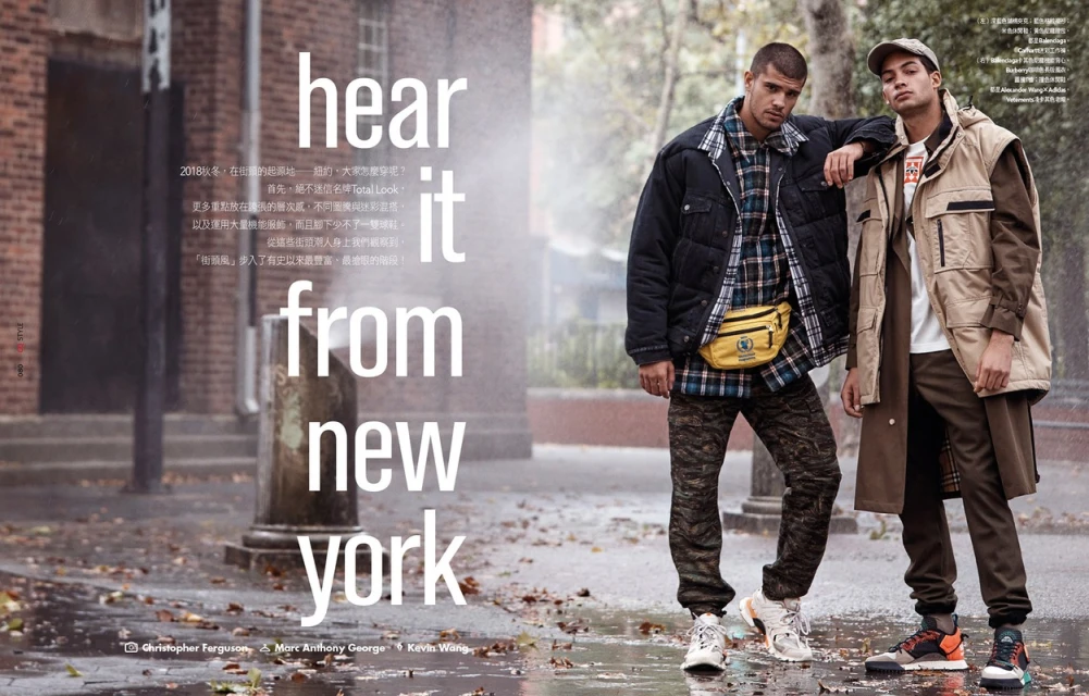 Two adult men pose on a wet city street in New York, wearing fashionable outerwear and sneakers. The backdrop features urban architecture.