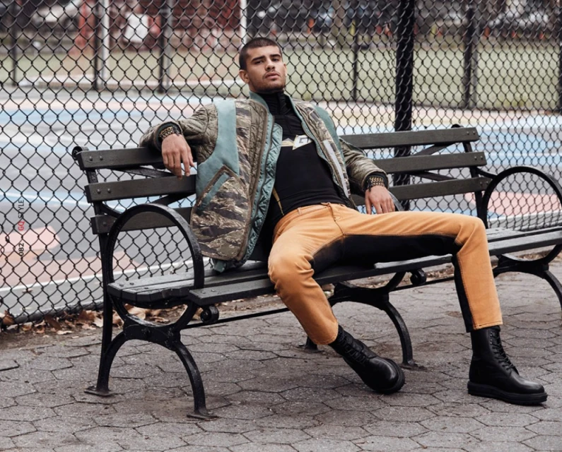 Man in jacket and orange pants rests on a black park bench outdoors with gray patterned ground and a chain-link fence in the background.