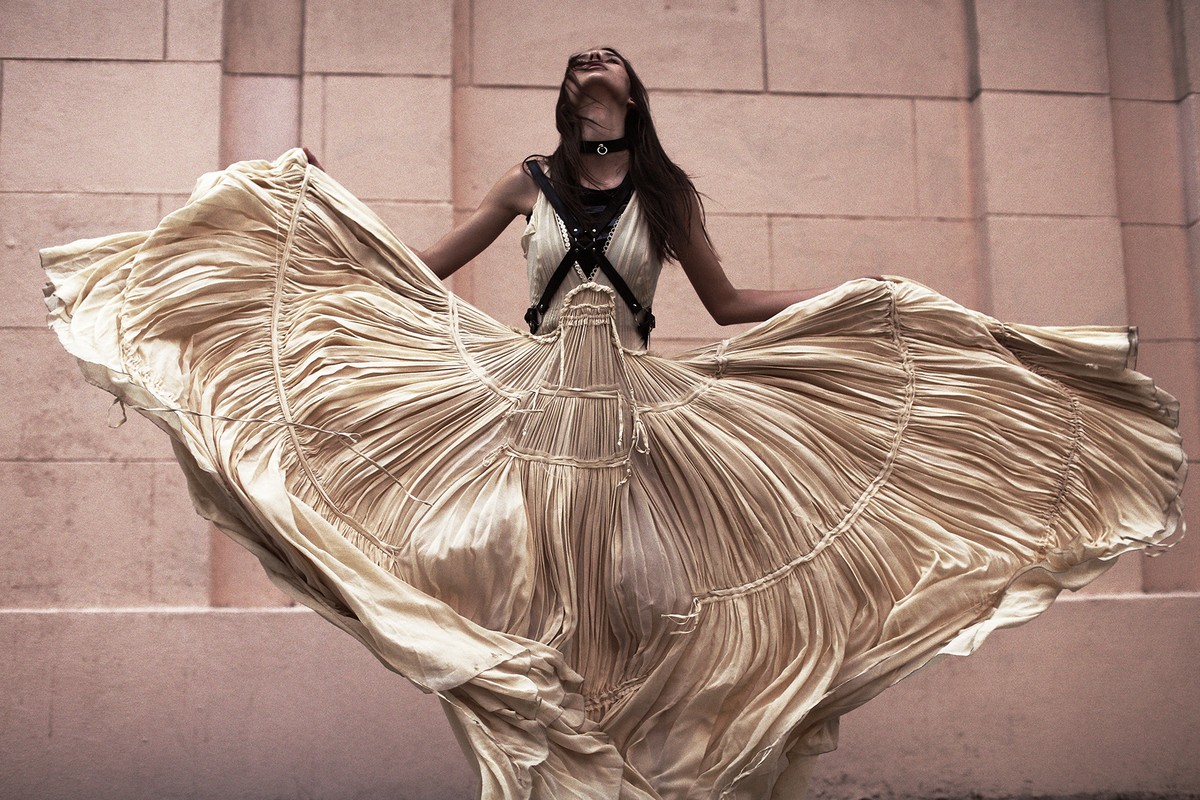 Woman in flowing gold dress dances with arms outstretched against a textured wall, fashion photography, neutral color palette.