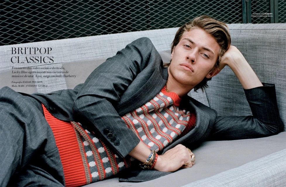 Blonde male model in gray suit and patterned knit shirt poses on gray couch in a fashion editorial shot against a dark grey background.