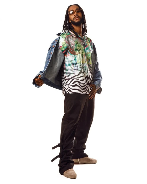 Studio shot of an adult male with dreadlocks, wearing a patterned shirt, denim jacket, and dark pants against a white backdrop.
