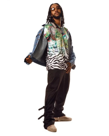Studio shot of an adult male with dreadlocks, wearing a patterned shirt, denim jacket, and dark pants against a white backdrop.