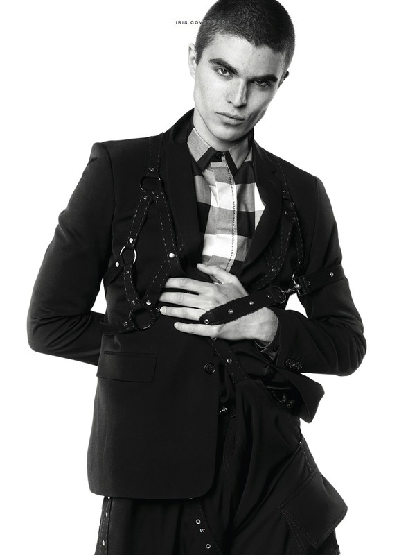 Monochrome fashion portrait of a male model in a black blazer, harness, and checkered shirt, against a white backdrop.