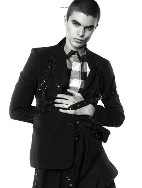 Monochrome fashion portrait of a male model in a black blazer, harness, and checkered shirt, against a white backdrop.