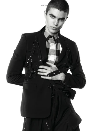 Monochrome fashion portrait of a male model in a black blazer, harness, and checkered shirt, against a white backdrop.