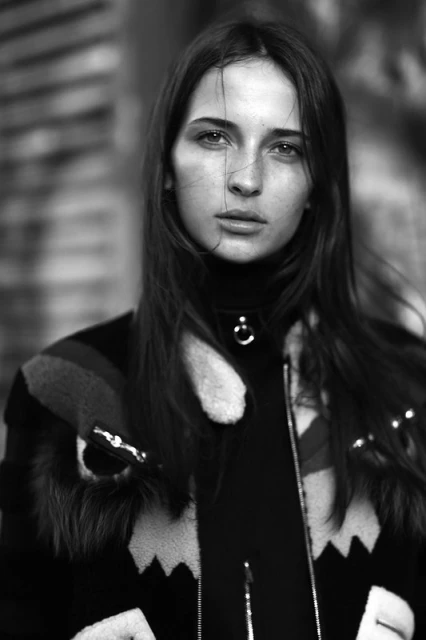 Black and white portrait of a brunette woman with long hair, wearing a turtleneck and patterned jacket, against an indistinct background.