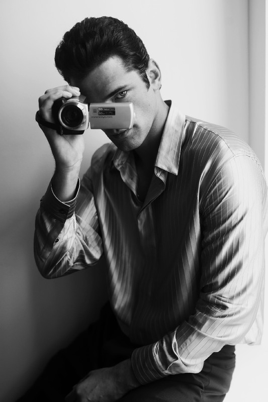 Black and white studio portrait of a brunette adult male holding a handheld camcorder up to his eye, looking directly at the viewer.